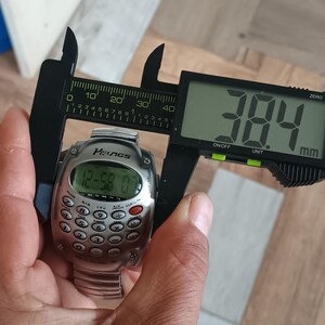 May include: A silver digital watch with a calculator keypad is measured with a digital caliper. The watch face displays the time. The caliper reads 38.4 mm, approximately 1.5 inches.