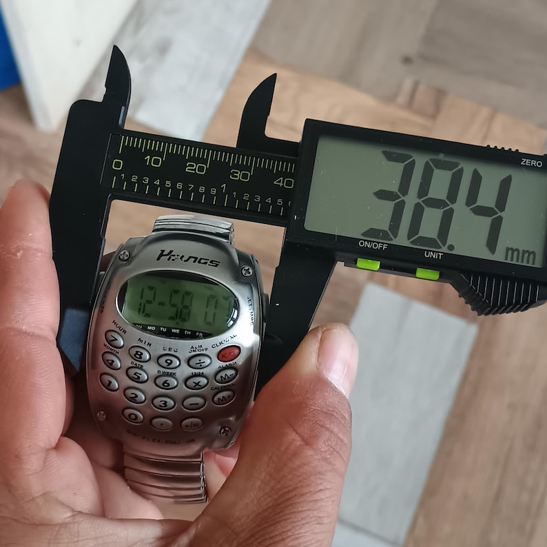 May include: A silver digital watch with a calculator keypad is measured with a digital caliper. The watch face displays the time. The caliper reads 38.4 mm, approximately 1.5 inches.