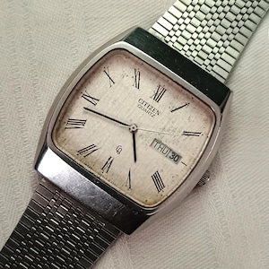 Könnte beinhalten: Eine Vintage Citizen Quartz-Uhr mit einem silberfarbenen, rechteckigen Gehäuse und einem Metallarmband. Das Zifferblatt ist cremefarben mit schwarzen römischen Ziffern und Zeigern. Der Tag wird als "DO 30" angezeigt.
