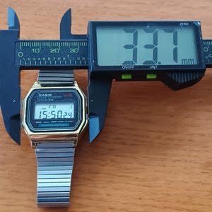 May include: A gold-toned Casio digital watch with a silver band is measured with a black digital caliper. The watch face displays the time as 15:50:24. The caliper reads 33.1 mm.