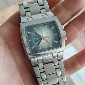 May include: A silver-toned wristwatch with a square face and a metallic band. The watch face has a light-colored center with dark blue and gold accents. The watch is displayed on a person's hand.