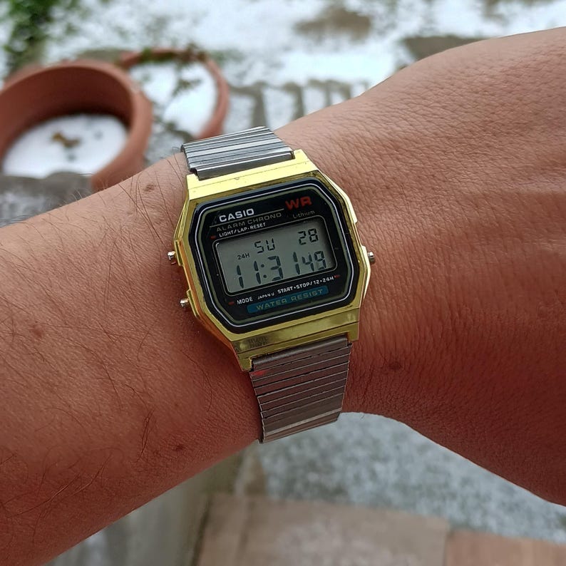 May include: A gold-toned Casio digital watch with a silver band is displayed. The watch face shows the time as 11:31:49, with the day and date visible. The watch has the text "CASIO" and "WATER RESIST" on the face.