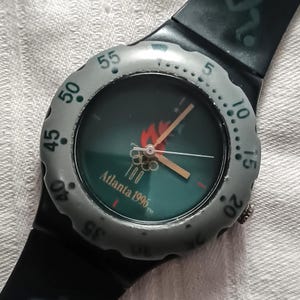 Puede incluir: Reloj de pulsera negro con esfera verde con el logotipo de los Juegos Olímpicos de Atlanta 1996. El reloj tiene un bisel gris con números blancos y una correa negra. Las manecillas del reloj son doradas.