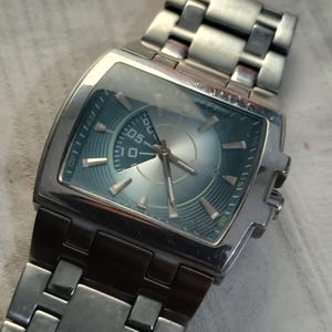 May include: Silver-toned wristwatch with a rectangular face and a blue-green dial. The watch features a stainless steel band and a digital display showing the number "05". The watch has a modern, industrial design.