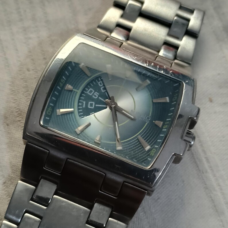 May include: Silver-toned wristwatch with a rectangular face and a blue-green dial. The watch features a stainless steel band and a digital display showing the number "05". The watch has a modern, industrial design.