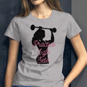 Könnte beinhalten: Graues T-Shirt mit einer schwarzen Silhouette einer Frau, die eine Langhantel hebt. Der Text "Practice Safe Sets" ist in Pink unter der Silhouette geschrieben.
