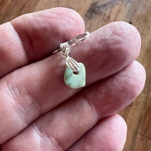 Peut inclure: Une boucle d'oreille en argent avec un petit pendentif en pierre vert clair de forme irr&eacute;guli&egrave;re. La pierre a un trou pr&egrave;s du sommet et est attach&eacute;e &agrave; un cercle en argent. La boucle d'oreille est tenue dans une main.