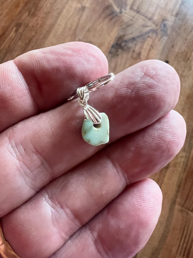 Peut inclure: Une boucle d'oreille en argent avec un petit pendentif en pierre vert clair de forme irr&eacute;guli&egrave;re. La pierre a un trou pr&egrave;s du sommet et est attach&eacute;e &agrave; un cercle en argent. La boucle d'oreille est tenue dans une main.