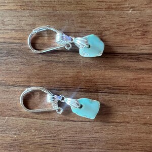 Peut inclure: Une paire de boucles d'oreilles &agrave; crochet argent&eacute;es avec des pendentifs en pierre turquoise de forme irr&eacute;guli&egrave;re. Les boucles d'oreilles sont pr&eacute;sent&eacute;es sur une surface en bois, mettant en valeur leur design et leur contraste de couleurs.