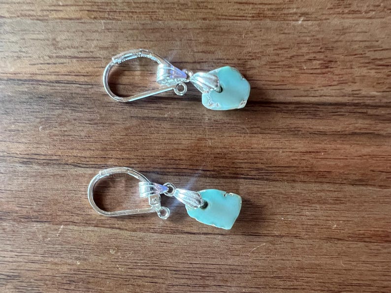 Peut inclure: Une paire de boucles d'oreilles &agrave; crochet argent&eacute;es avec des pendentifs en pierre turquoise de forme irr&eacute;guli&egrave;re. Les boucles d'oreilles sont pr&eacute;sent&eacute;es sur une surface en bois, mettant en valeur leur design et leur contraste de couleurs.