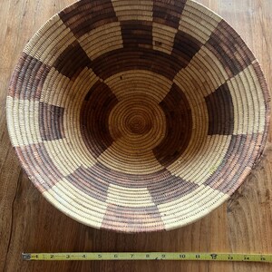 May include: A round woven basket with a geometric pattern in shades of brown and beige. The basket is approximately 15 inches in diameter and has a shallow depth. The design features concentric circles and rectangular shapes.