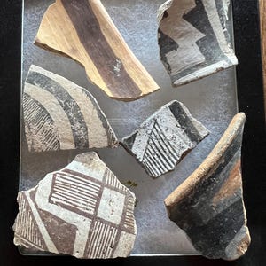 May include: A collection of ancient pottery shards with geometric designs in black, brown, and white. The fragments are displayed in a black shadow box, showcasing the intricate patterns and textures of the historical artifacts.