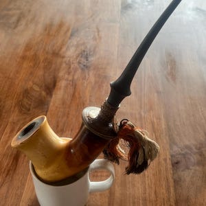 May include: A wooden smoking pipe with a brown and black stem and a white ceramic mug. The pipe is resting in the mug.