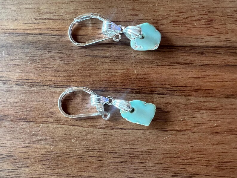 Peut inclure: Une paire de boucles d'oreilles &agrave; crochet argent&eacute;es avec des pendentifs en pierre turquoise de forme irr&eacute;guli&egrave;re. Les boucles d'oreilles sont pr&eacute;sent&eacute;es sur une surface en bois, mettant en valeur leur design et leur couleur.