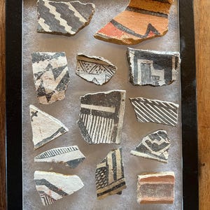 May include: A framed collection of ancient pottery shards. The fragments feature geometric designs in black, red, and white against a tan background. The pottery pieces are displayed in a black frame.