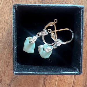 Peut inclure: Une paire de boucles d'oreilles avec des pierres bleu clair de forme irr&eacute;guli&egrave;re, serties dans du m&eacute;tal argent&eacute;. Les boucles d'oreilles sont pr&eacute;sent&eacute;es dans une petite bo&icirc;te noire, sur une surface en bois. Elles ont un fermoir &agrave; levier.