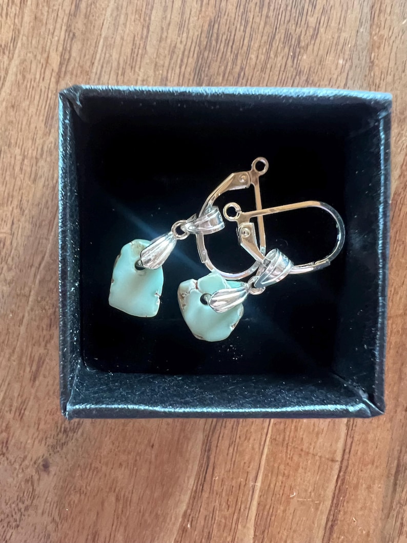 Peut inclure: Une paire de boucles d'oreilles avec des pierres bleu clair de forme irr&eacute;guli&egrave;re, serties dans du m&eacute;tal argent&eacute;. Les boucles d'oreilles sont pr&eacute;sent&eacute;es dans une petite bo&icirc;te noire, sur une surface en bois. Elles ont un fermoir &agrave; levier.