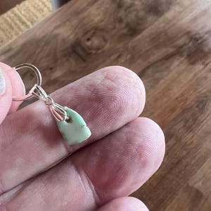 Peut inclure: Un petit pendentif vert clair avec une b&eacute;li&egrave;re argent&eacute;e. Le pendentif est de forme irr&eacute;guli&egrave;re, peut-&ecirc;tre une pierre naturelle. Le fond est une surface en bois.