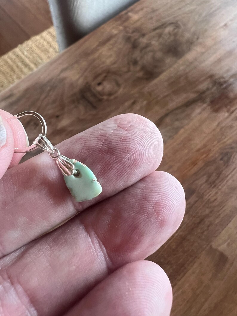 Peut inclure: Un petit pendentif vert clair avec une b&eacute;li&egrave;re argent&eacute;e. Le pendentif est de forme irr&eacute;guli&egrave;re, peut-&ecirc;tre une pierre naturelle. Le fond est une surface en bois.