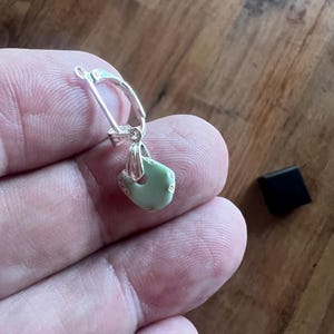 Peut inclure: Boucle d'oreille en argent avec fermoir &agrave; levier. La boucle d'oreille pr&eacute;sente un petit pendentif en pierre vert clair de forme irr&eacute;guli&egrave;re. Un petit carr&eacute; noir est en arri&egrave;re-plan.