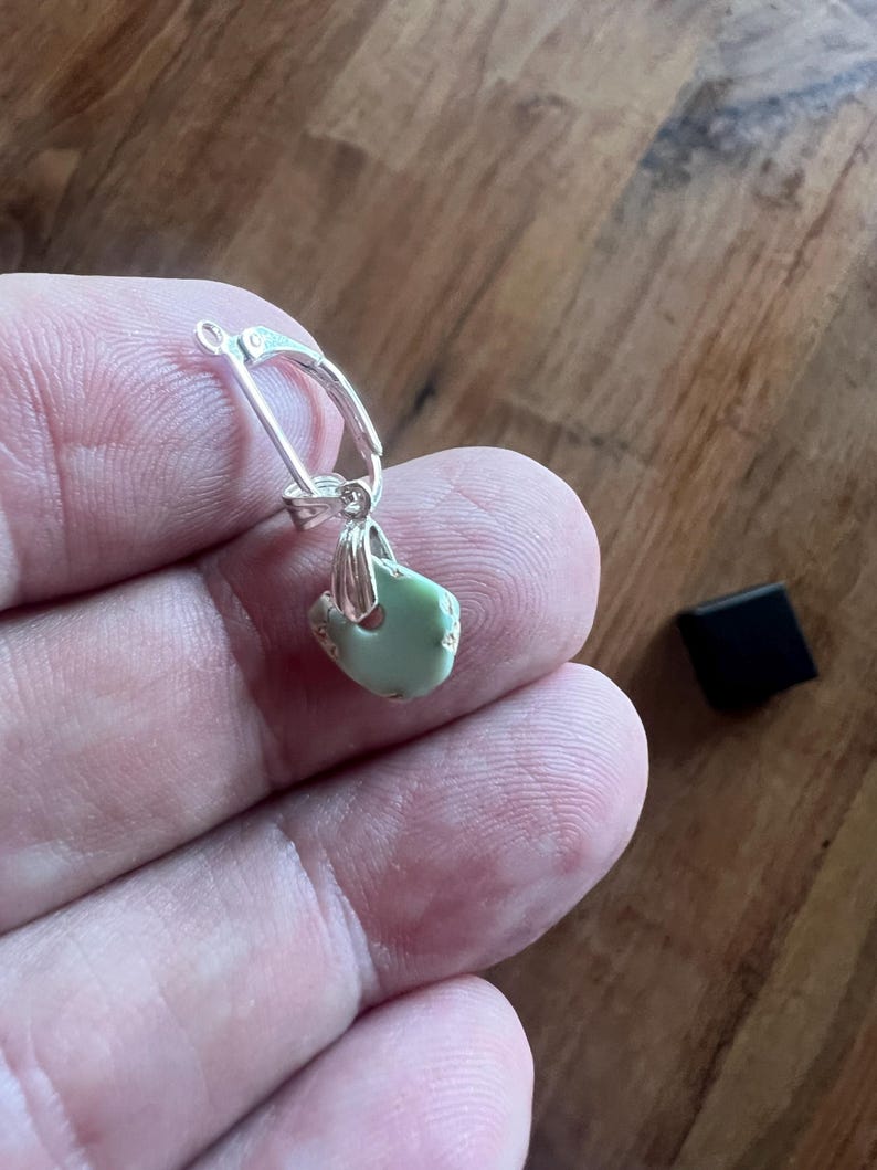 Peut inclure: Boucle d'oreille en argent avec fermoir &agrave; levier. La boucle d'oreille pr&eacute;sente un petit pendentif en pierre vert clair de forme irr&eacute;guli&egrave;re. Un petit carr&eacute; noir est en arri&egrave;re-plan.