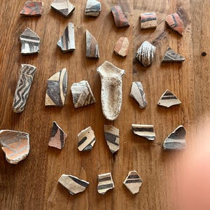 May include: A collection of ancient pottery shards with various geometric designs in black, red, and brown. The fragments are arranged on a wooden surface, showcasing the remnants of historical craftsmanship and artistry.