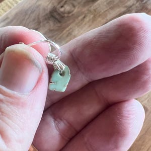 Peut inclure: Une boucle d'oreille unique avec un pendentif vert clair de forme irr&eacute;guli&egrave;re. Le pendentif est attach&eacute; &agrave; un cerceau de couleur argent&eacute;e. La boucle d'oreille est tenue dans la main d'une personne sur un fond en bois.