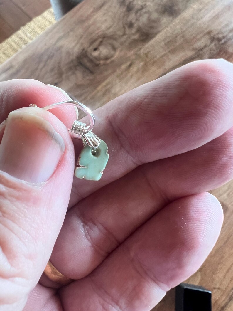 Peut inclure: Une boucle d'oreille unique avec un pendentif vert clair de forme irr&eacute;guli&egrave;re. Le pendentif est attach&eacute; &agrave; un cerceau de couleur argent&eacute;e. La boucle d'oreille est tenue dans la main d'une personne sur un fond en bois.