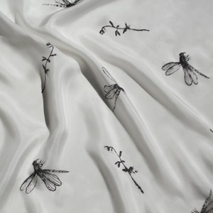 May include: White fabric featuring a repeating pattern of black dragonflies and sprigs. The fabric is draped, showing soft folds and a silky sheen. The dragonfly illustrations vary in size and orientation, creating a dynamic design.