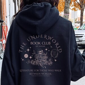 Puede incluir: Sudadera con capucha negra con un diseño gráfico en la espalda. El diseño presenta el texto "The Underworld Book Club" con una ilustración de libros, estrellas y elementos botánicos. La sudadera también tiene el texto "Literature for those who walk between worlds."
