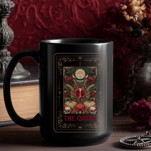 Puede incluir: Taza de cer&aacute;mica negra con dise&ntilde;o de carta del tarot que presenta una granada, calaveras y el texto "THE QUEEN". La taza tiene un asa grande y un acabado brillante.