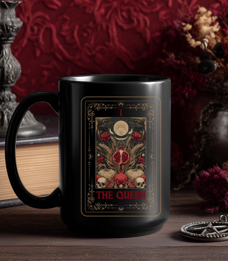 Puede incluir: Taza de cer&aacute;mica negra con dise&ntilde;o de carta del tarot que presenta una granada, calaveras y el texto "THE QUEEN". La taza tiene un asa grande y un acabado brillante.