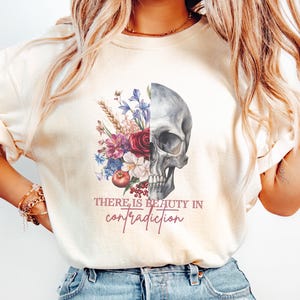 Puede incluir: Una camiseta blanca con un diseño floral y un cráneo. El texto "There is beauty in contradiction" está impreso en la camiseta.