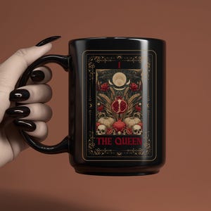 Puede incluir: Taza de cer&aacute;mica negra con dise&ntilde;o de carta del tarot. La carta presenta una granada, calaveras y elementos florales, con las palabras "THE QUEEN" en dorado. La taza tiene un asa curva y un acabado brillante.