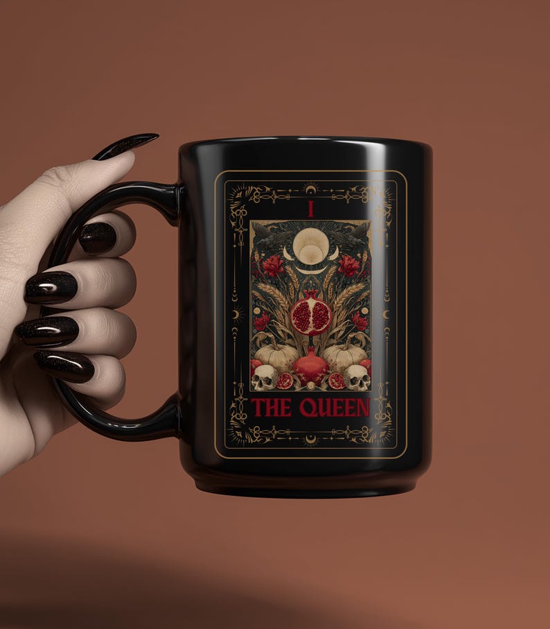 Puede incluir: Taza de cer&aacute;mica negra con dise&ntilde;o de carta del tarot. La carta presenta una granada, calaveras y elementos florales, con las palabras "THE QUEEN" en dorado. La taza tiene un asa curva y un acabado brillante.