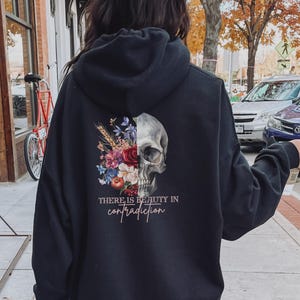 Puede incluir: Sudadera con capucha negra con un diseño de calavera y flores en la espalda. El texto "THERE IS BEAUTY IN CONTRADICTION" está impreso debajo del diseño.