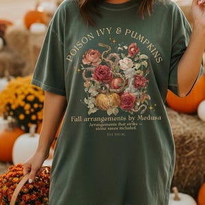 May include: Dark green t-shirt with the text "POISON IVY & PUMPKINS" and "Fall arrangements by Medusa." The shirt features a floral and pumpkin design. The shirt also has the text "Arrangements that strike - stone vases included. EST 700 BC."