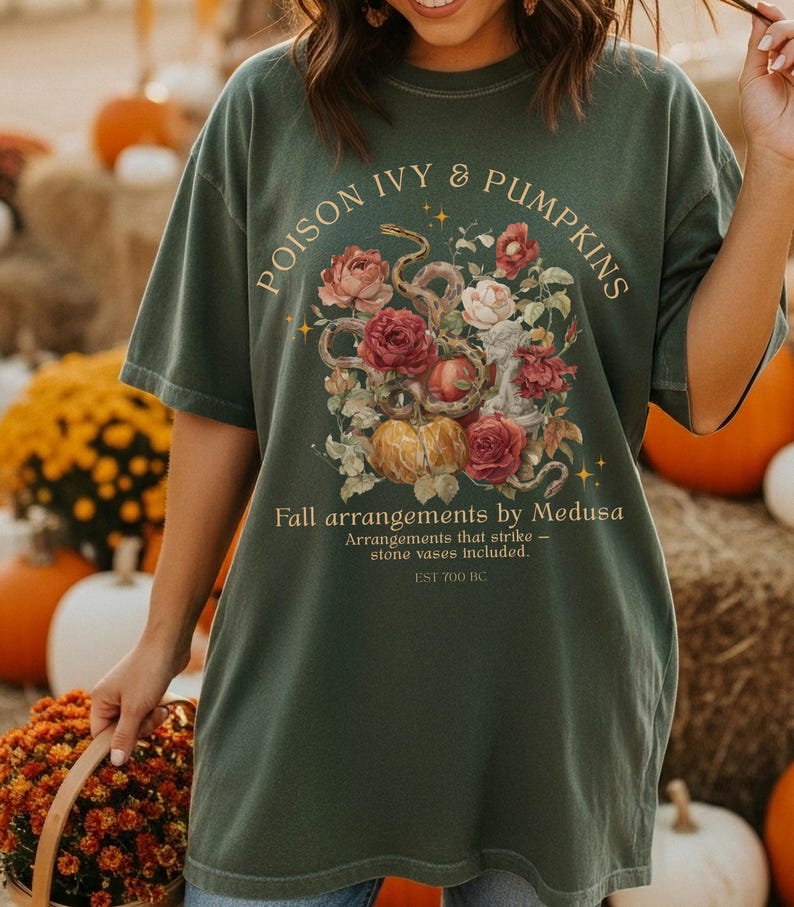 May include: Dark green t-shirt with the text "POISON IVY & PUMPKINS" and "Fall arrangements by Medusa." The shirt features a floral and pumpkin design. The shirt also has the text "Arrangements that strike - stone vases included. EST 700 BC."