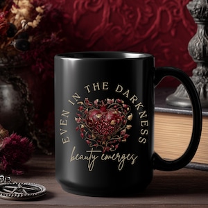 Op de afbeelding: Zwarte keramische mok met een hartvormig bloemmotief in rood en goud. De mok heeft de tekst "EVEN IN THE DARKNESS beauty emerges" in gouden letters. De mok heeft een groot oor en staat op een houten oppervlak.