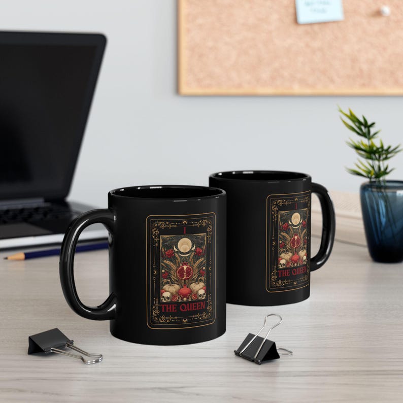 Puede incluir: Dos tazas de cer&aacute;mica negra con un dise&ntilde;o de carta del tarot. La carta presenta una imagen central de una granada, calaveras y el texto "THE QUEEN". El dise&ntilde;o est&aacute; enmarcado en dorado sobre un fondo oscuro.
