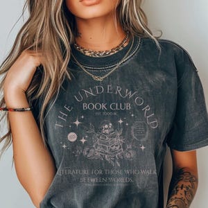 Puede incluir: Una camiseta negra con un diseño gráfico de estilo vintage que presenta una calavera sosteniendo una pila de libros. El texto dice "The Underworld Book Club Est. 3000 BC" y "Literatura para aquellos que caminan entre mundos."