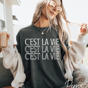 May include: A dark gray sweatshirt with the phrase "C'est la vie" repeated three times in white outlined text.