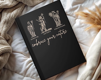 May include: A black journal with gold illustrations of Demeter, Artemis, and Persephone. The words "embrace your nature" are written in gold script below the illustrations. The journal is resting on a white and beige textured surface.