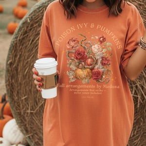 May include: Orange t-shirt with the text "POISON IVY & PUMPKINS" and a floral and snake graphic. The shirt also has the text "Fall arrangements by Medusa" and "EST 700 BC". A person is holding a white coffee cup.