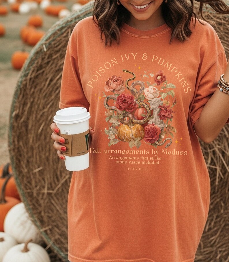May include: Orange t-shirt with the text "POISON IVY & PUMPKINS" and a floral and snake graphic. The shirt also has the text "Fall arrangements by Medusa" and "EST 700 BC". A person is holding a white coffee cup.