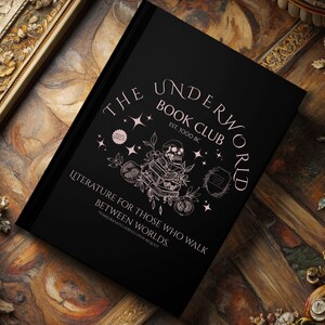 Puede incluir: Un libro negro con el título "The Underworld Book Club" en una fuente estilizada, con un diseño de calavera y flores. La portada también incluye la frase "Literature for Those Who Walk Between Worlds."