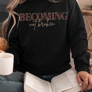 Może przedstawiać: Czarna bluza z napisem "BECOMING not broken" w kwiatowy wzór. Bluza ma okrągły dekolt i długie rękawy.