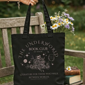Puede incluir: Bolso tote negro con el texto "THE UNDERWORLD BOOK CLUB" y un diseño de calavera. La bolsa está llena de flores y la sostiene una persona. El texto "LITERATURE FOR THOSE WHO WALK BETWEEN WORLDS" también está impreso en la bolsa.