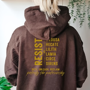May include: Brown hooded sweatshirt with the text "RESIST" in gold lettering. The text is vertically aligned and includes the names of mythological figures: Medusa, Hecate, Lilith, Lamia, Circe, and Sirens. Below the names is the text "DEFY. UNLEASH. RECLAIM petrify the patriarchy".