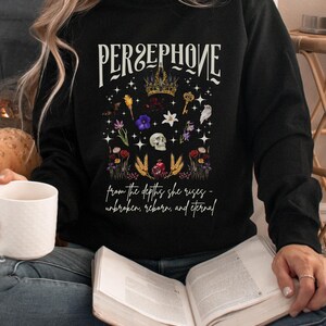 Puede incluir: Sudadera negra con un diseño gráfico que presenta el nombre "Persephone" en texto blanco con una corona y elementos florales. El texto debajo dice "from the depths she rises - unbroken, reborn, and eternal."